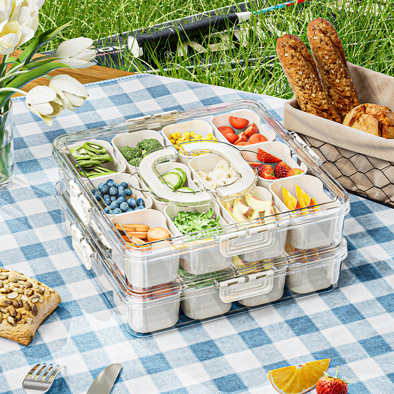 Clear plastic food container with compartments filled with snacks on a picnic setup with bread and flowers.