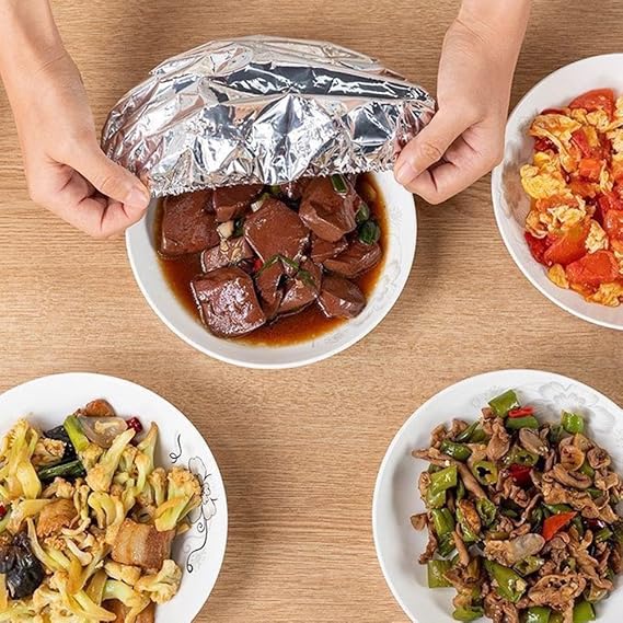 Person opening a foil-wrapped dish on a wooden table with various other dishes around.