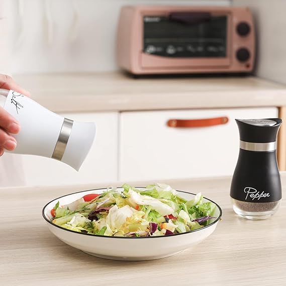 Person seasoning a salad with a white pepper grinder, with a black pepper grinder and bowl of salad in the foreground.