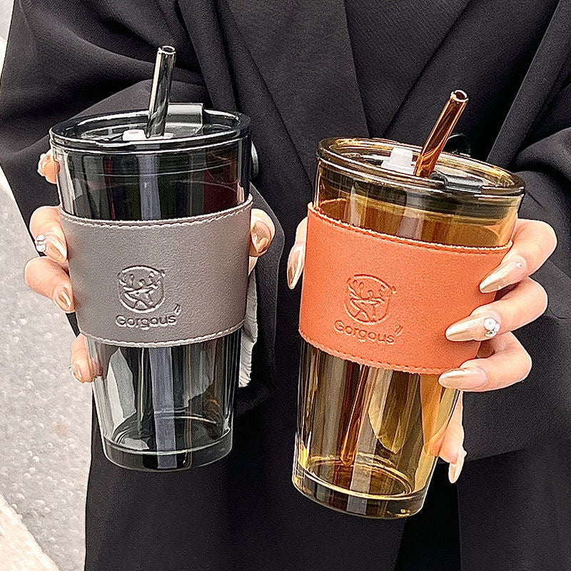 Two tumblers with straws held by a person, one in gray sleeve and one in brown sleeve, on a neutral background.