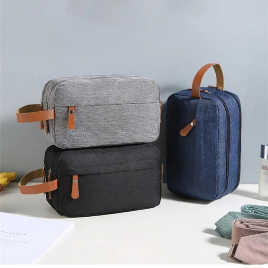 Three toiletry bags in gray, black, and blue with brown straps on a light surface.