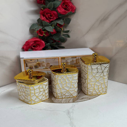 Set of three decorative canisters with gold lids on a marble surface with flowers in the background.