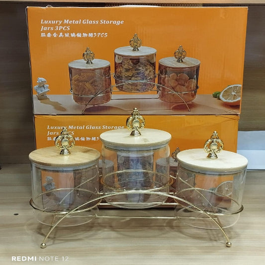 Set of metal and glass storage jars with a decorative stand on a wooden surface, with packaging in the background.